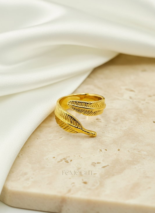 Gold feather-shaped ring on a textured beige surface with a white fabric background
