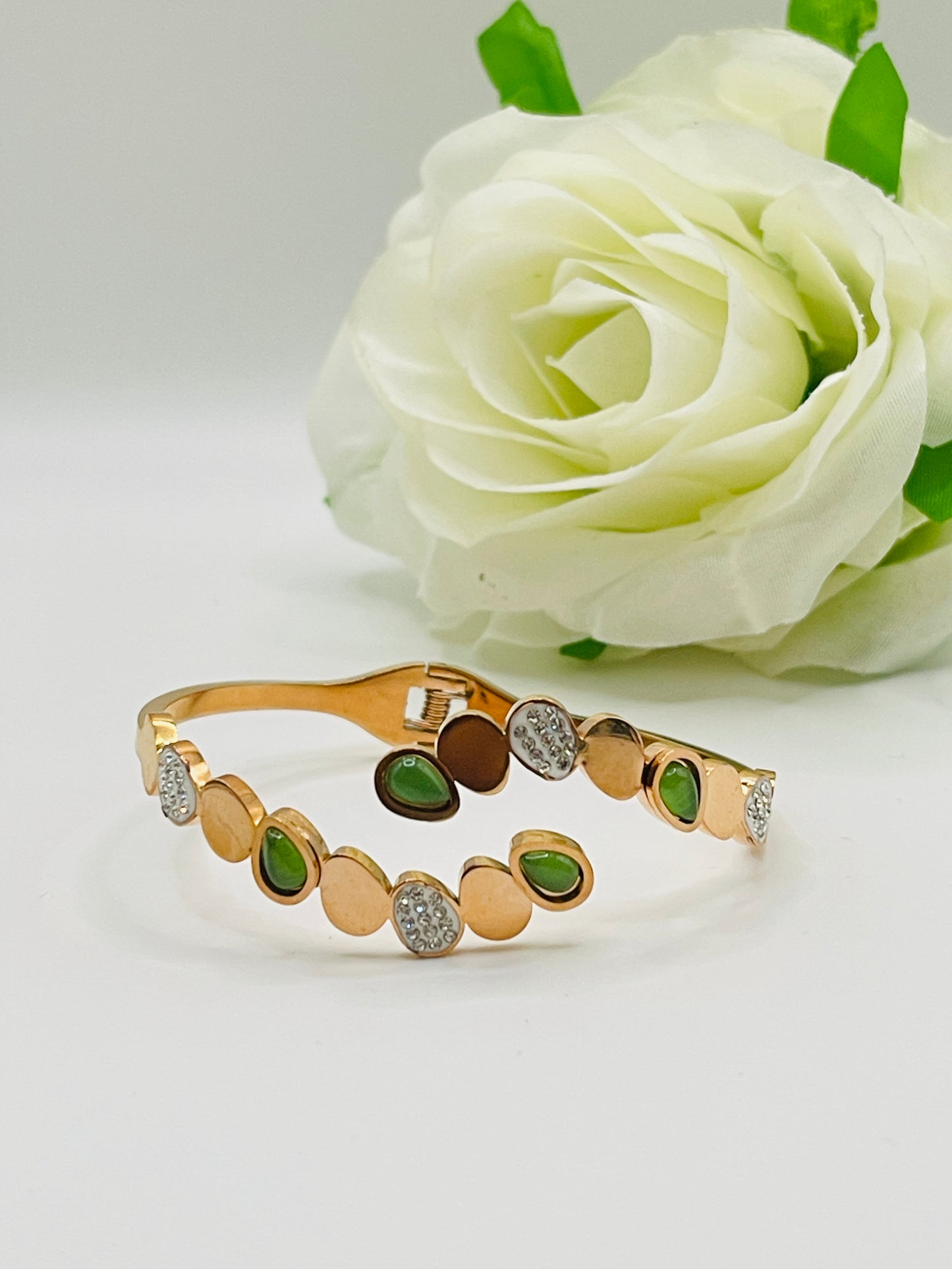 Rose Gold bracelet with green stones and diamonds in front of a white rose on a light gray background