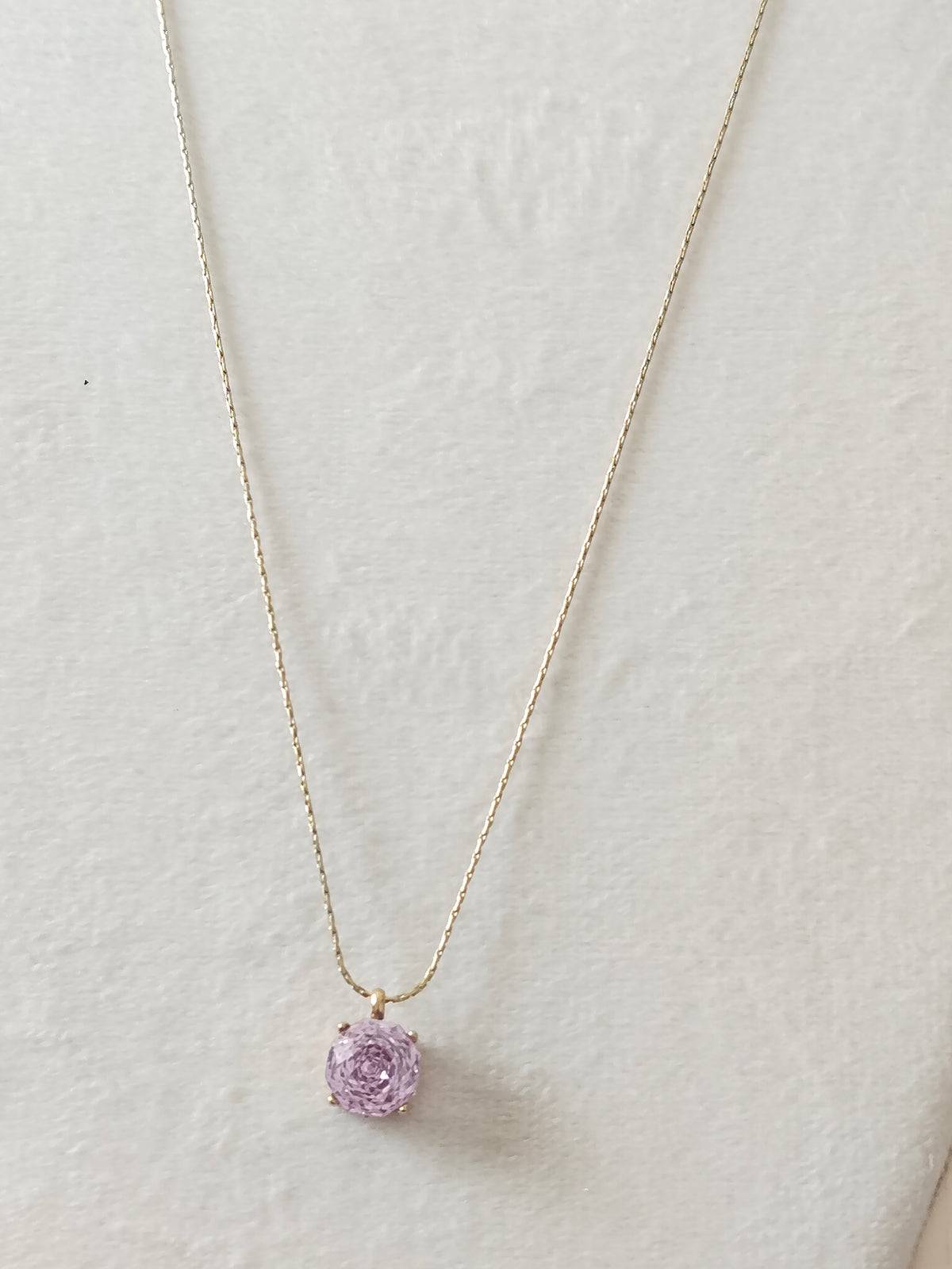 A delicate gold chain necklace with a small, round, light purple rose-shaped gemstone pendant, displayed on a textured off-white background.