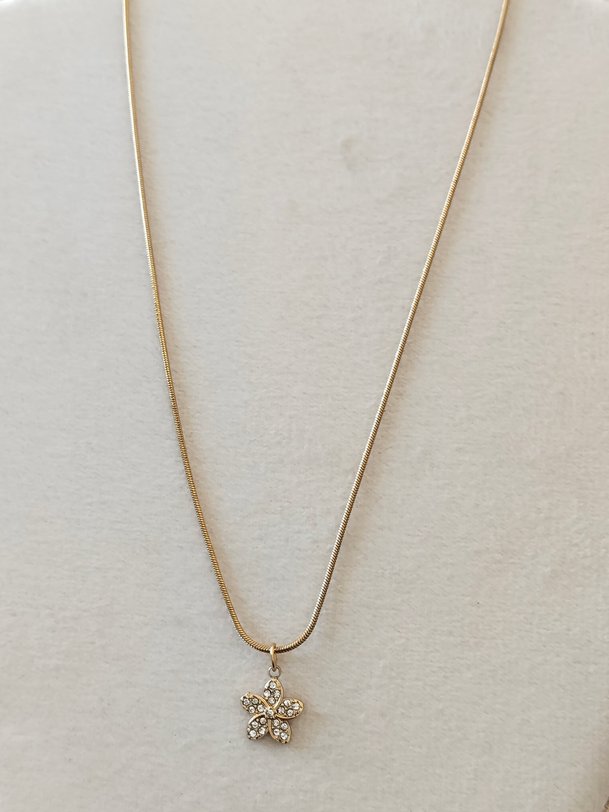 A delicate gold snake chain necklace with a small flower-shaped pendant set with clear rhinestones, displayed on a textured beige surface.