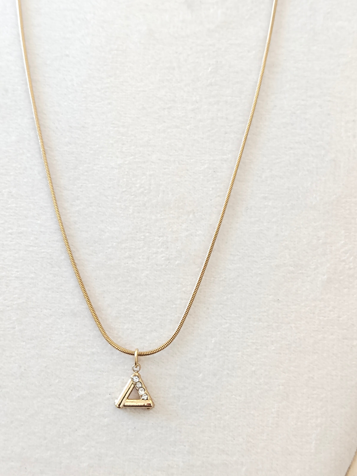 A delicate gold chain necklace with a small triangular pendant featuring a tiny clear stone, displayed on a textured off-white jewelry bust.