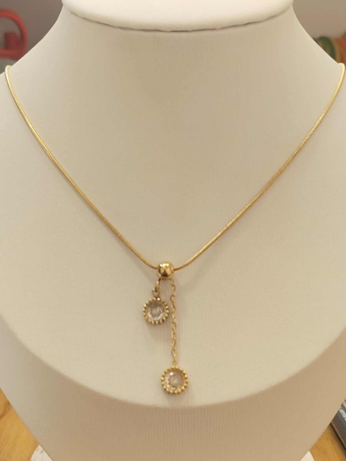 A delicate gold chain necklace displayed on a cream-colored jewelry bust. The necklace features a small gold clasp with two dangling circular charms, each adorned with a clear stone surrounded by a gold textured border.