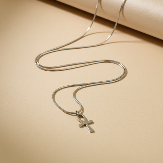 A silver snake chain necklace with a small cross pendant, displayed on a warm beige background with a rolled cream paper at the top.