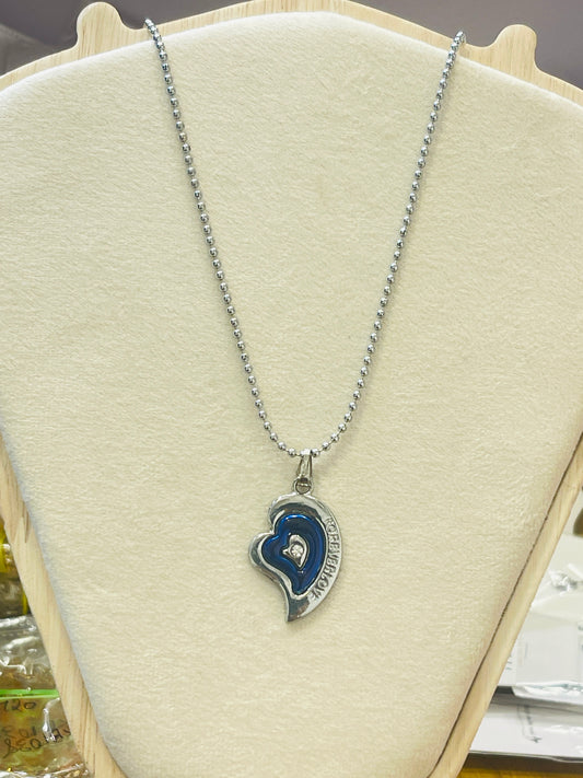 A silver ball-chain necklace with a heart-shaped pendant featuring a blue enamel inlay and a small clear center stone, displayed on a cream-colored jewelry bust.