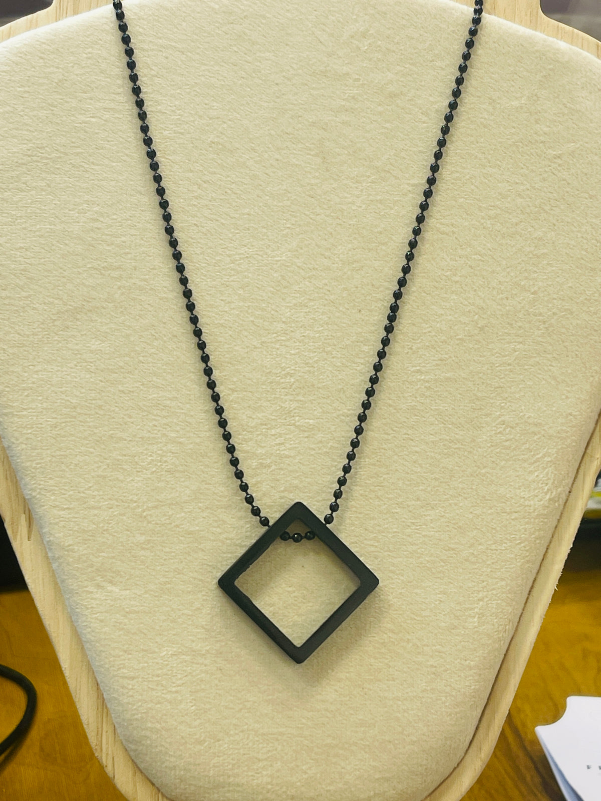 A black diamond-shaped pendant necklace with a black beaded chain, displayed on a cream-colored jewelry bust.