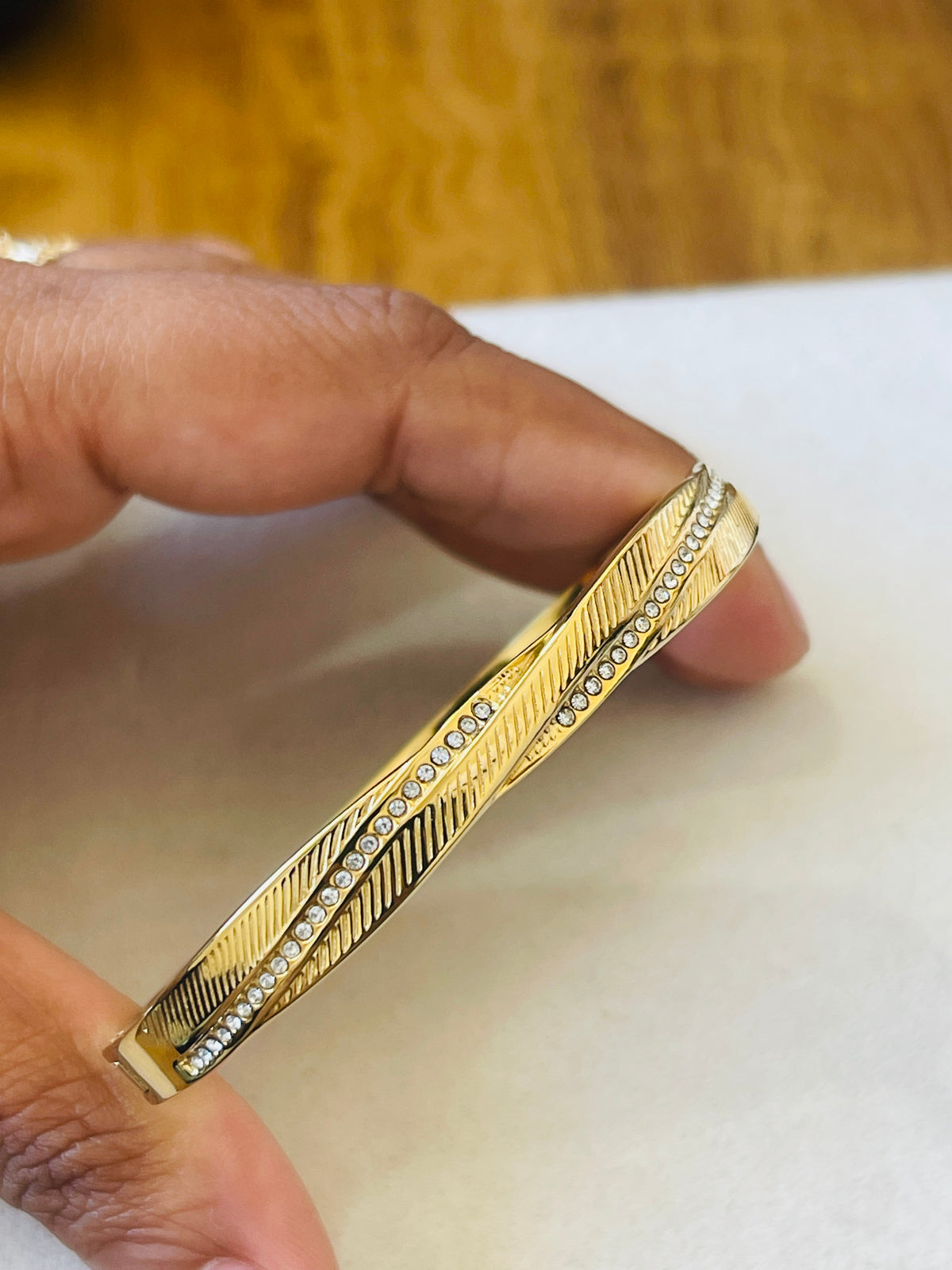A gold-toned bangle bracelet with a textured chevron pattern and a row of small clear stones, held between fingers against a white surface with a blurred wooden background.