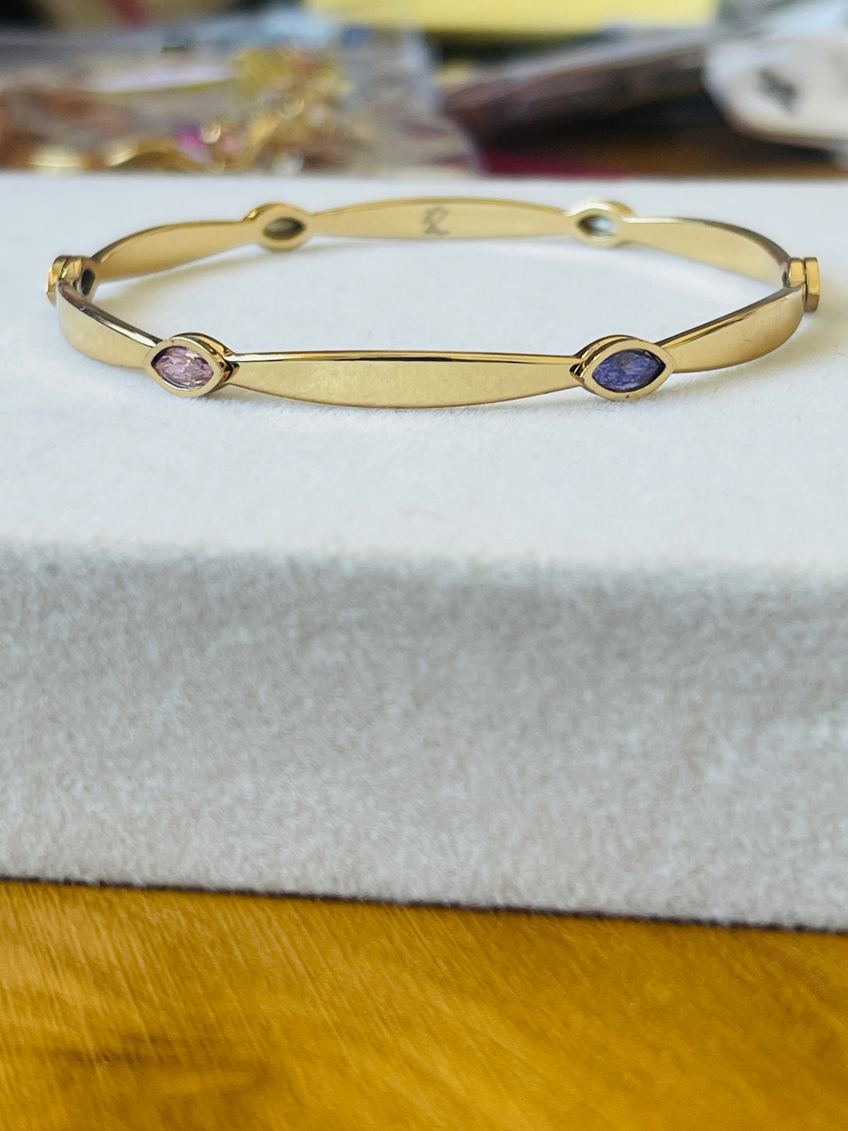 A gold-plated bangle bracelet with two small oval gemstone accents, one pinkish-purple and one dark blue, resting on a white textured jewelry display block.