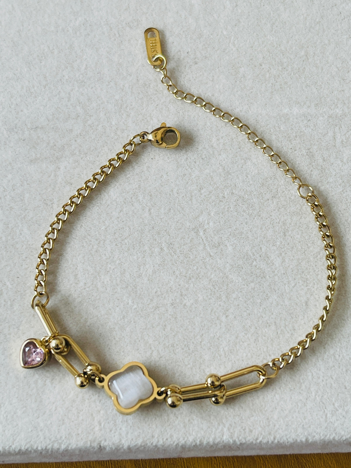 A gold chain bracelet with a four-leaf clover charm, a pink heart charm, and a white oval charm, displayed on a textured white surface.