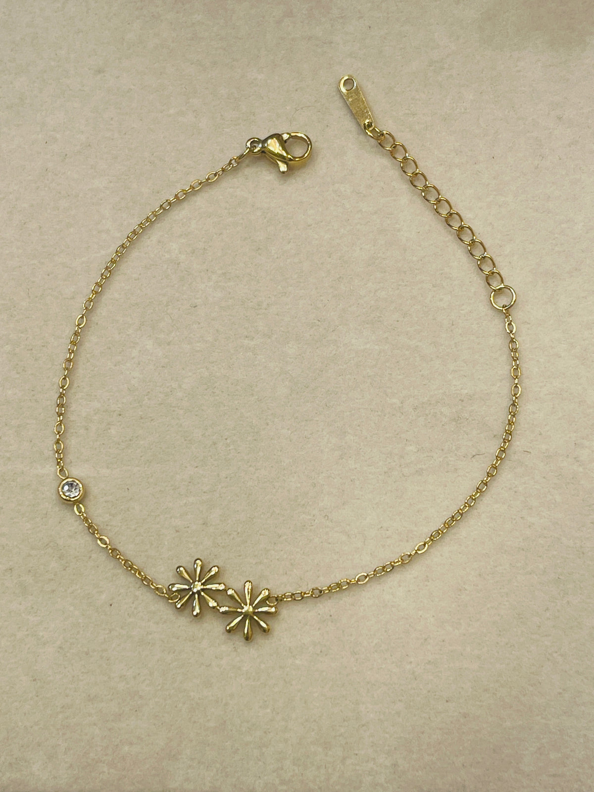 A delicate gold chain bracelet with two small starburst charms and a single clear rhinestone accent, displayed on a textured beige background.