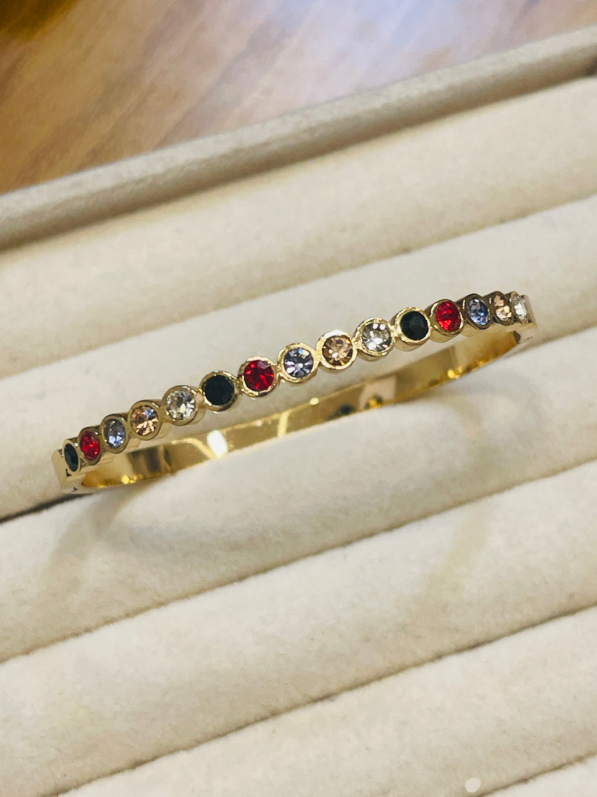A gold bangle bracelet with a row of colorful round gemstones, displayed on a beige jewelry tray.