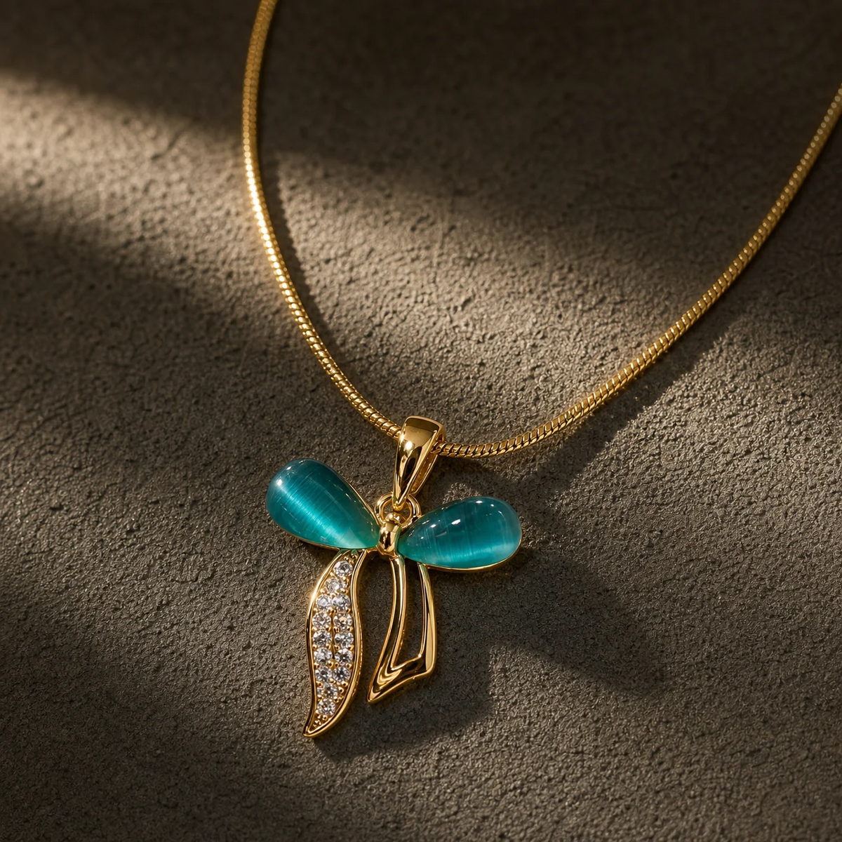 Gold pendant necklace with a bow-shaped design, featuring two glossy turquoise-blue cabochon stones and a leaf-like drop paved with small clear stones, displayed on a textured dark gray surface.
