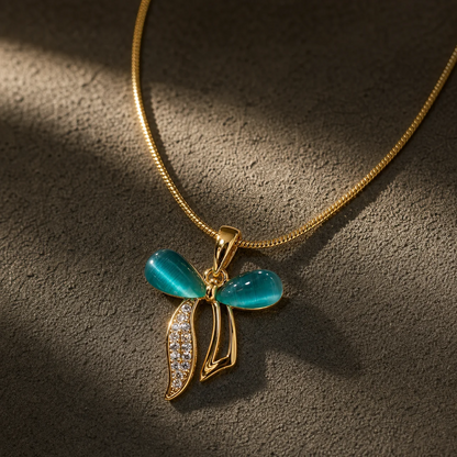 Gold pendant necklace with a bow-shaped design, featuring two glossy turquoise-blue cabochon stones and a leaf-like drop paved with small clear stones, displayed on a textured dark gray surface.