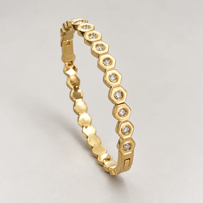 A gold bangle bracelet featuring a row of hexagonal links, each set with a small clear round gemstone, displayed on a neutral light gray background.