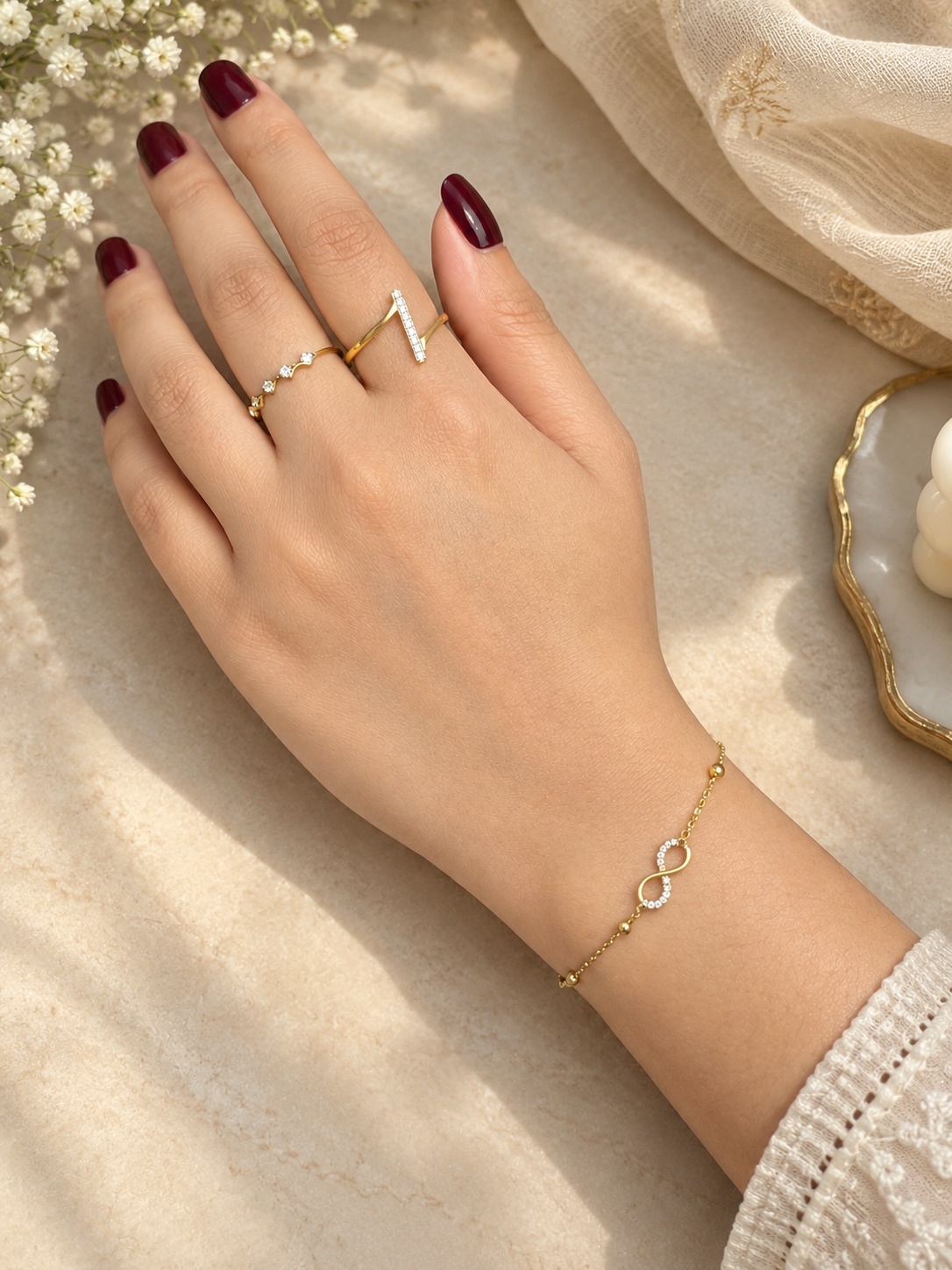 A close-up of a hand with dark burgundy nail polish, wearing a delicate gold chain bracelet with an infinity symbol and two gold rings, styled on a beige textured surface with dried flowers.