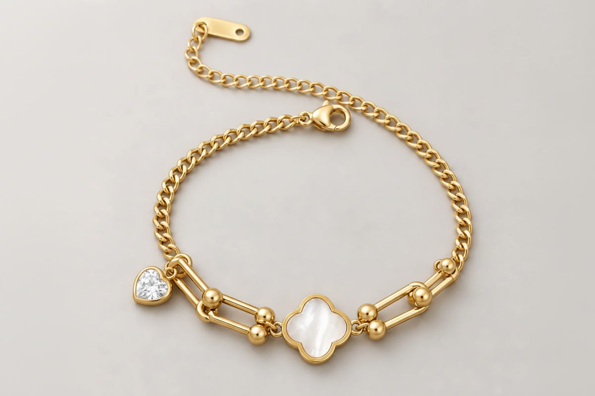 Gold chain bracelet with a four-leaf clover charm, a white heart-shaped crystal charm, and gold spacer beads, displayed on a neutral gray background.