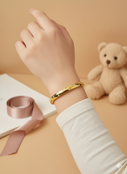 Hand wearing a gold bracelet with a teddy bear and ribbon in the background