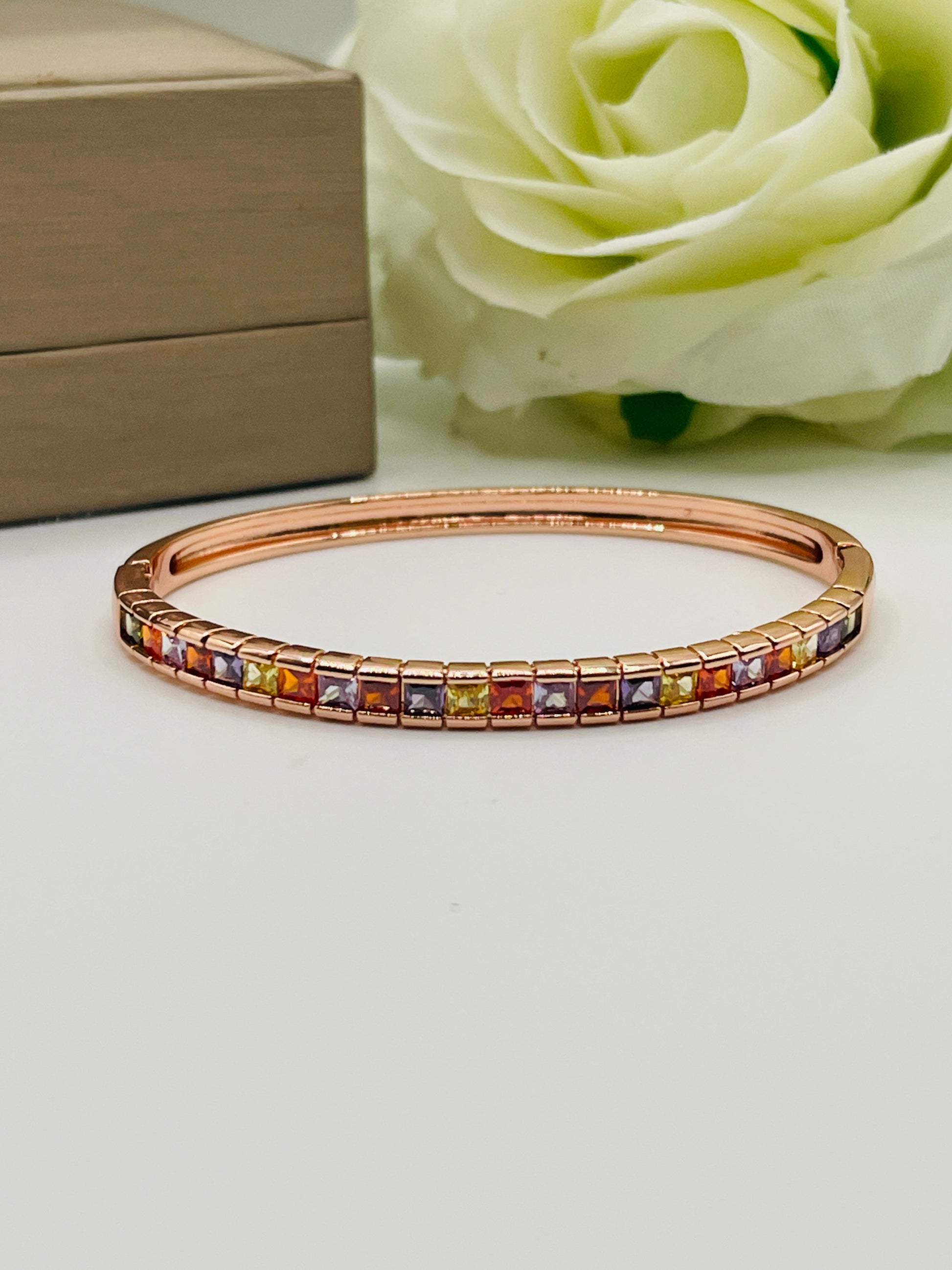 Rose gold bracelet with multicolored stones on a light surface with a white rose and box in the background.