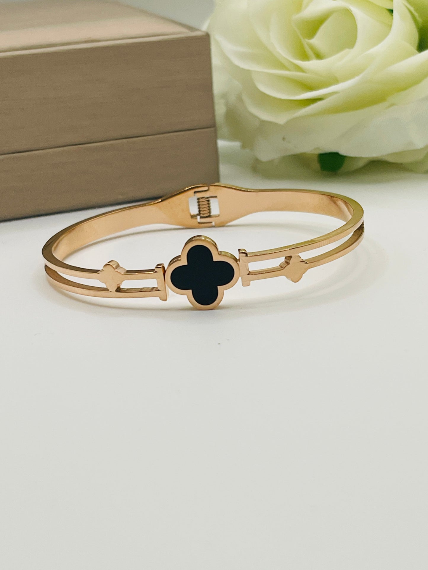 Gold bracelet with a black clover design on a white surface with a beige box and white rose in the background.