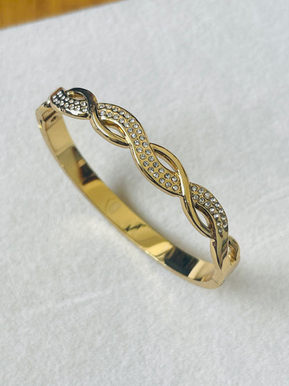 A gold bangle bracelet featuring an infinity-style link design embellished with small clear pavé stones, displayed on a textured white surface.