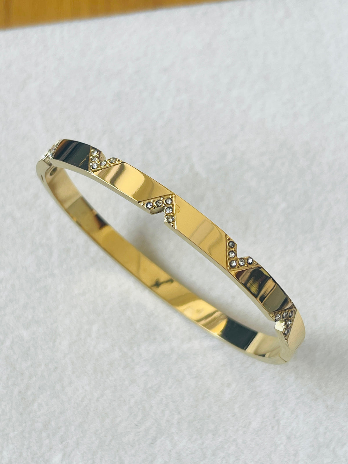 A polished gold bangle bracelet with small clear rhinestone accents arranged in diagonal lines, displayed on a textured white surface.