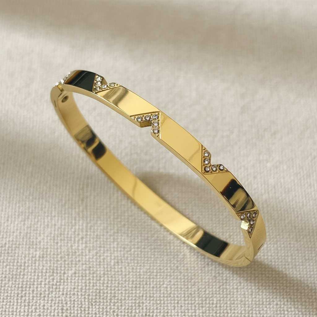 A polished gold bangle bracelet with geometric triangular inlays set with small clear stones, displayed on a textured beige fabric background.
