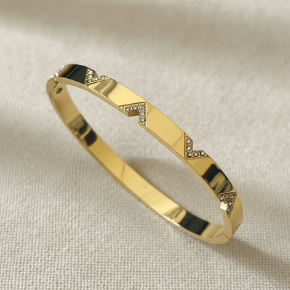 A polished gold bangle bracelet with geometric triangular inlays set with small clear stones, displayed on a textured beige fabric background.