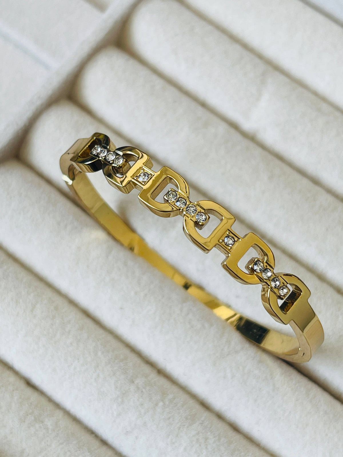A gold bangle bracelet with a repeating interlocking chain-link design, each accented with small clear stones, displayed on a white textured jewelry box.
