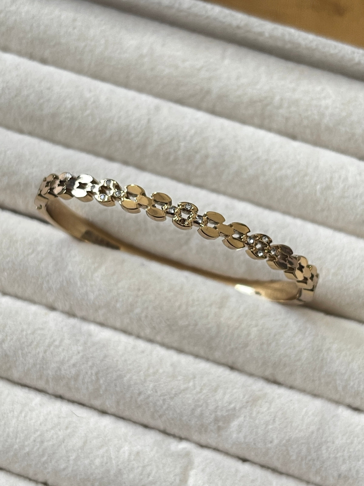 A delicate gold bangle bracelet with a row of small clear stones, displayed on a white textured ring box.
