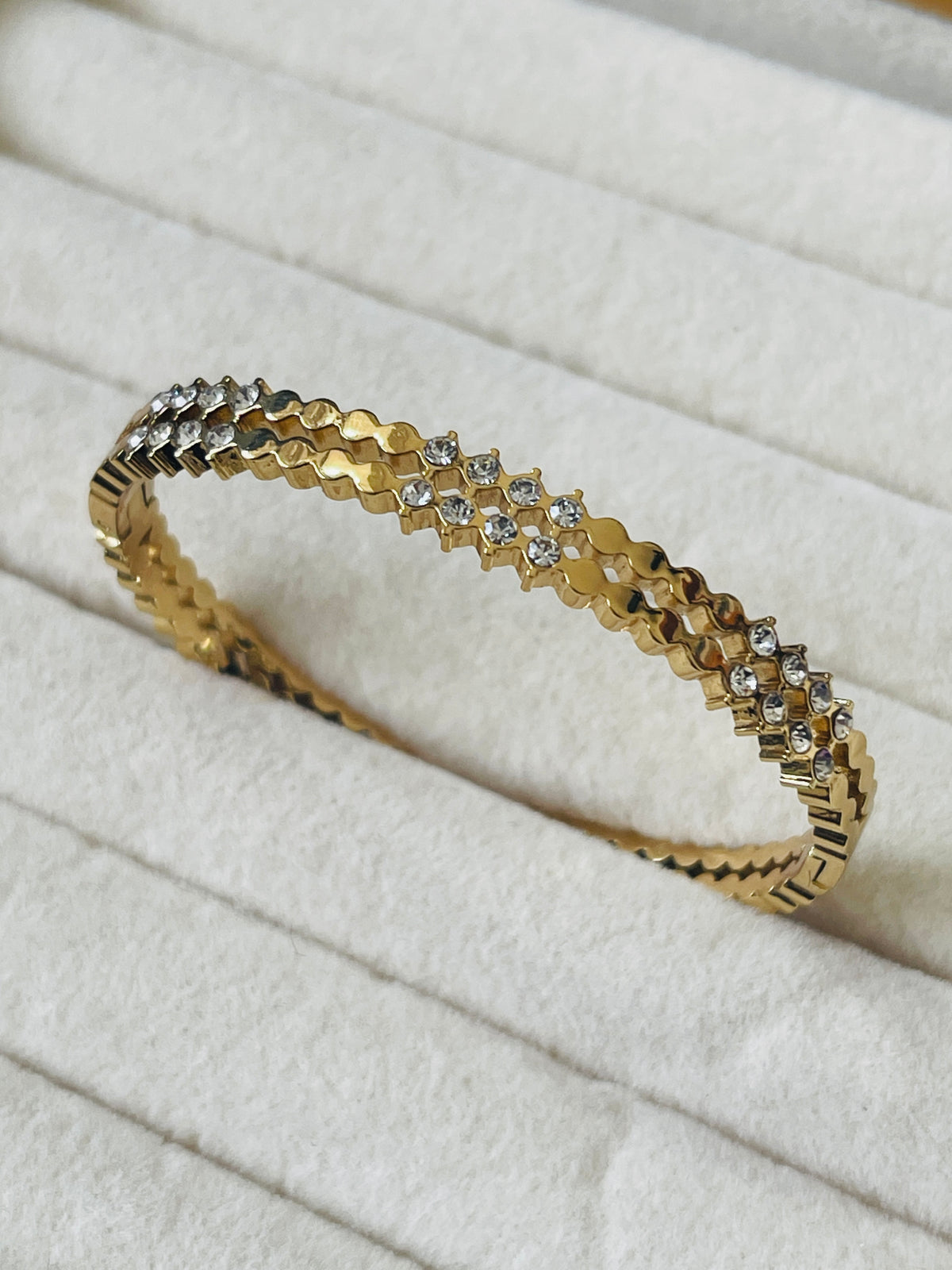 A gold bangle bracelet with a textured geometric pattern and small clear stones, displayed on a white textured fabric surface.