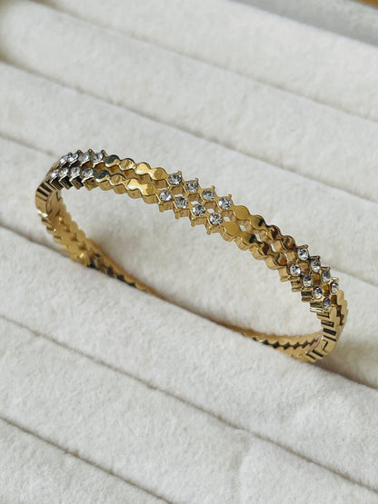 A gold bangle bracelet with a textured geometric pattern and small clear stones, displayed on a white textured fabric surface.