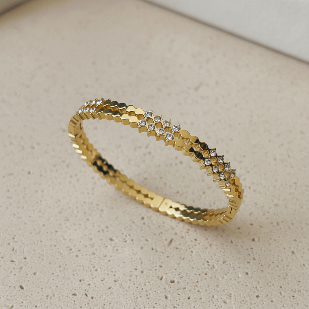 A gold-toned bangle bracelet with a textured geometric pattern and a row of small clear stones, resting on a light beige speckled surface.