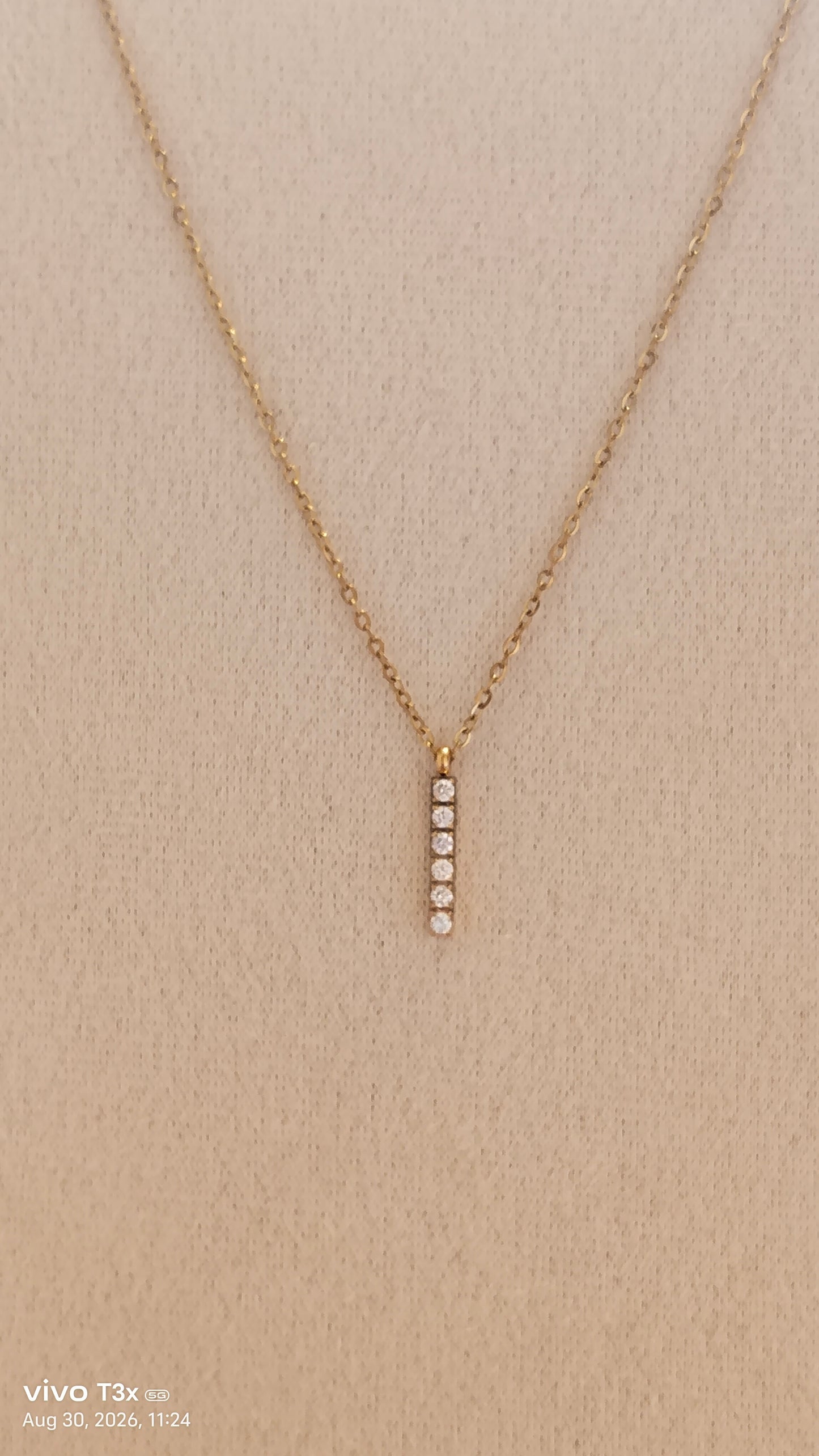 A delicate gold chain necklace with a vertical bar pendant featuring five small clear stones, displayed on a textured beige fabric background.