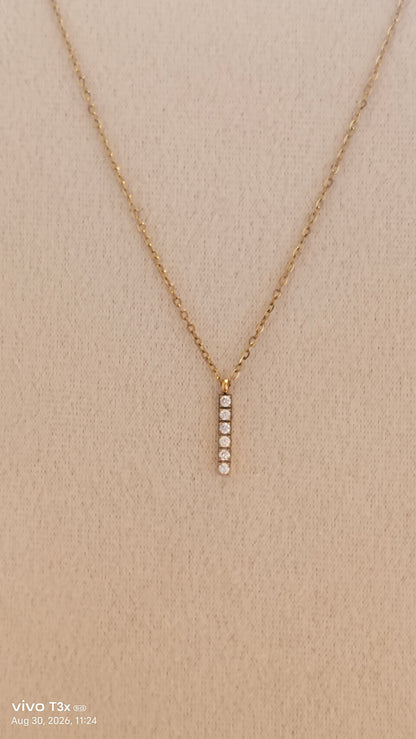 A delicate gold chain necklace with a vertical bar pendant featuring five small clear stones, displayed on a textured beige fabric background.