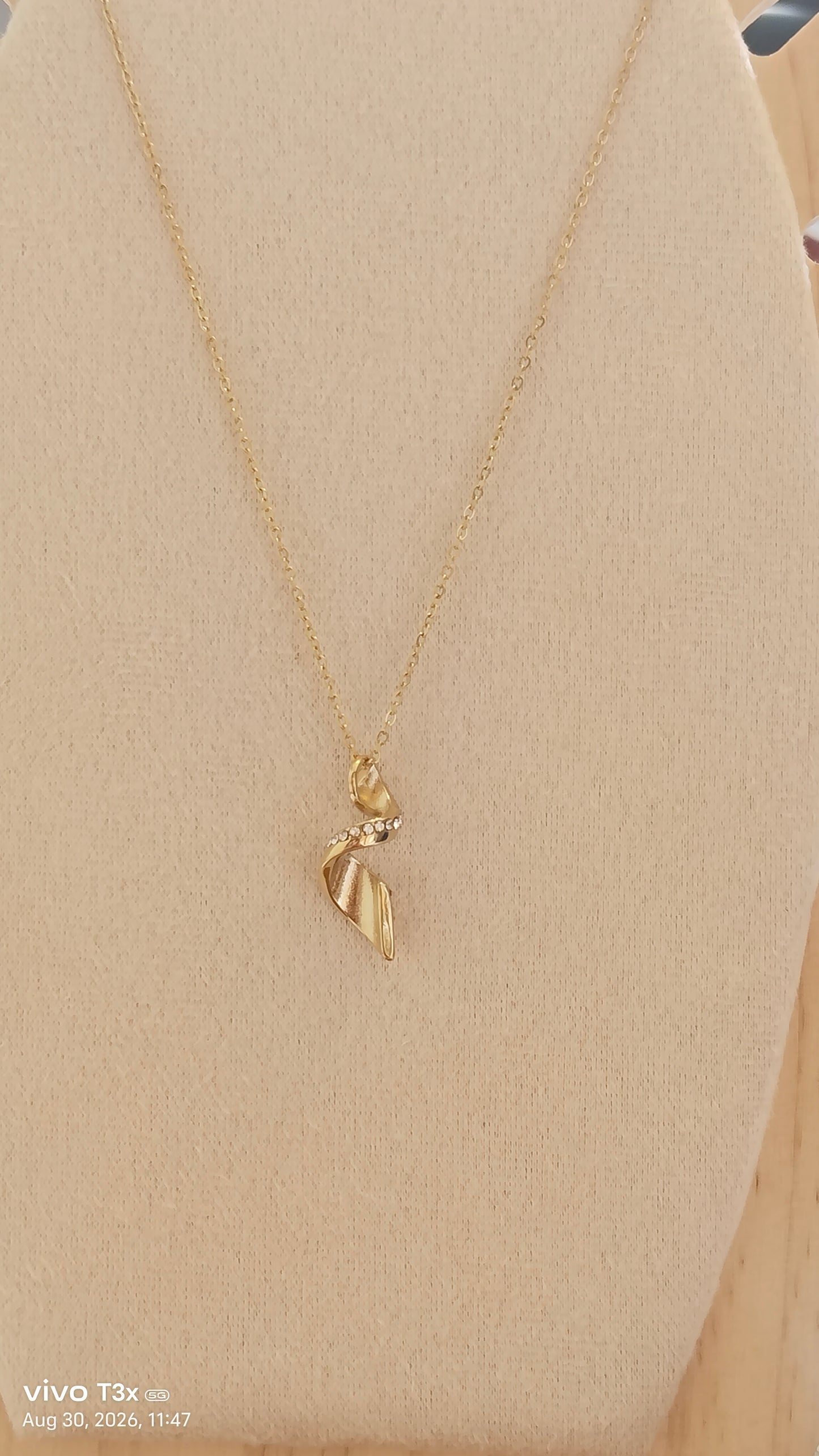 A delicate gold chain necklace with a small leaf-shaped pendant displayed on a beige fabric jewelry bust.