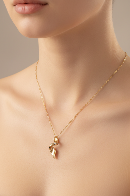A close-up of a model wearing a delicate gold chain necklace with a small ribbon-shaped pendant and a tiny pearl accent, set against a soft beige background.