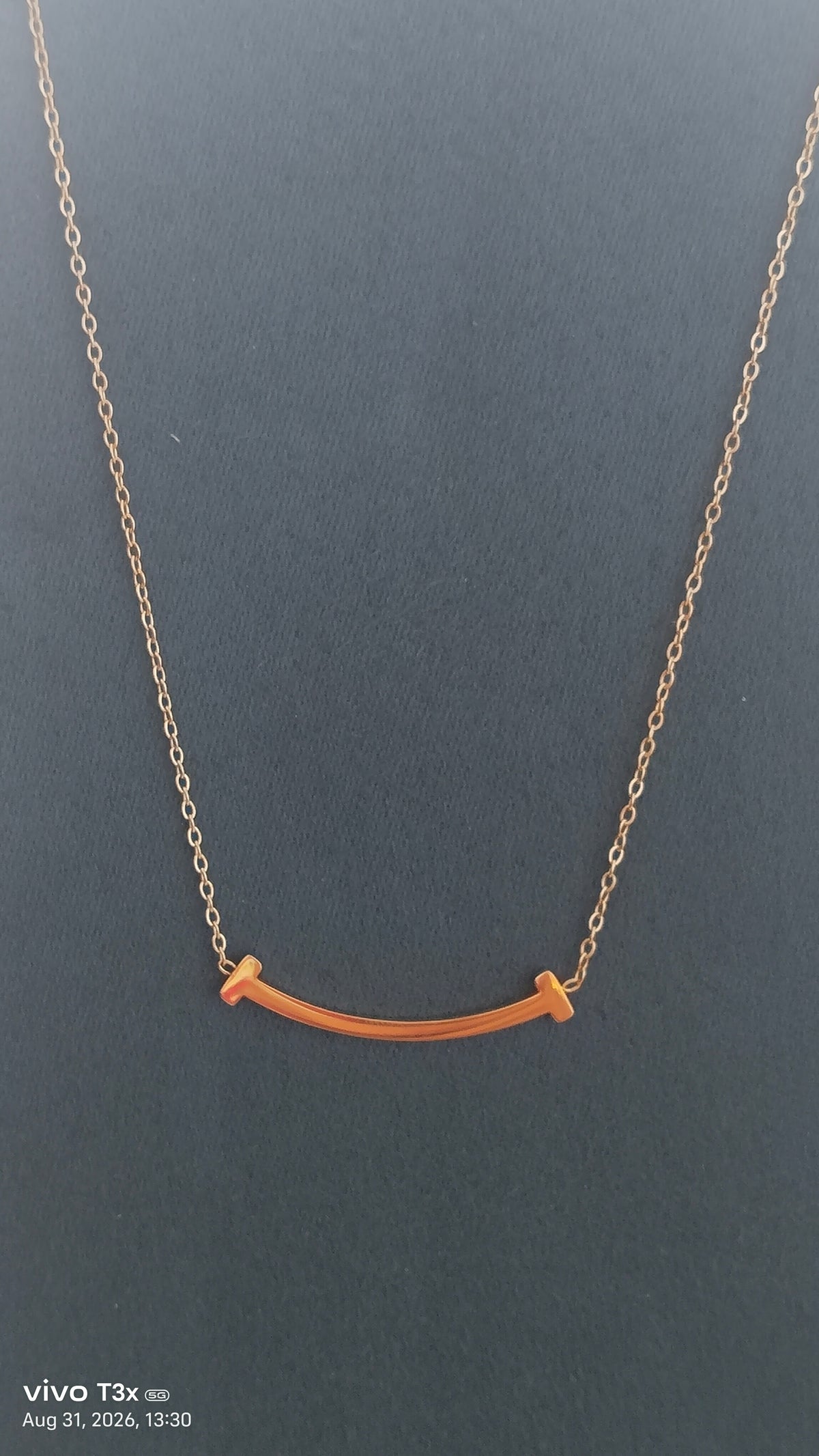 A delicate gold chain necklace featuring a small, curved bar pendant with two small, flat gold end-tips, displayed against a dark gray background.