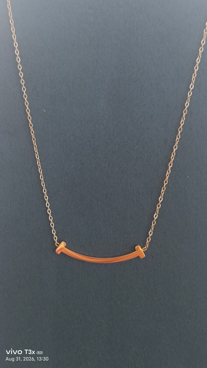 A delicate gold chain necklace featuring a small, curved bar pendant with two small, flat gold end-tips, displayed against a dark gray background.