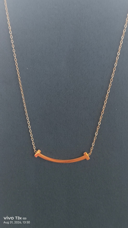 A delicate gold chain necklace featuring a small, curved bar pendant with two small, flat gold end-tips, displayed against a dark gray background.
