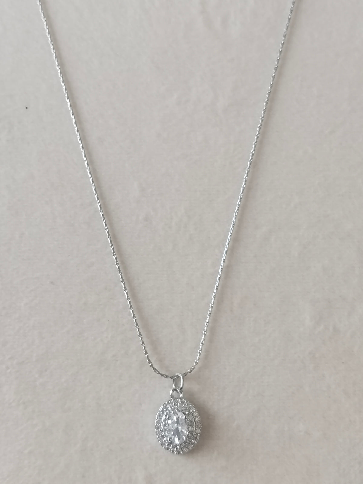 A silver teardrop pendant necklace with a pavé-set clear gemstone, displayed on a textured light beige background.