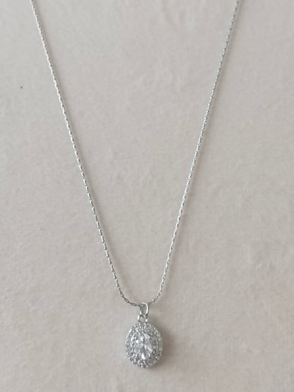A silver teardrop pendant necklace with a pavé-set clear gemstone, displayed on a textured light beige background.