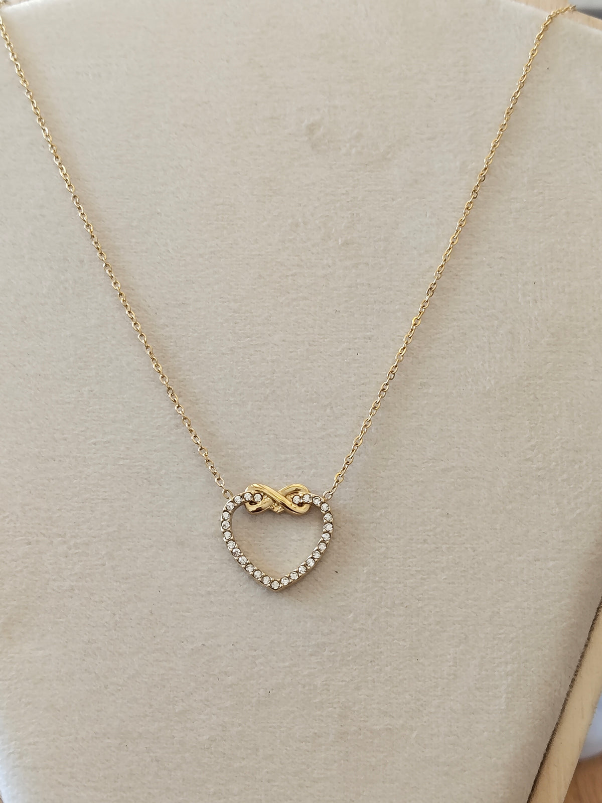 A delicate gold chain necklace with a heart-shaped pendant featuring a small infinity symbol at the top and a border of tiny clear stones, displayed on a beige jewelry bust.