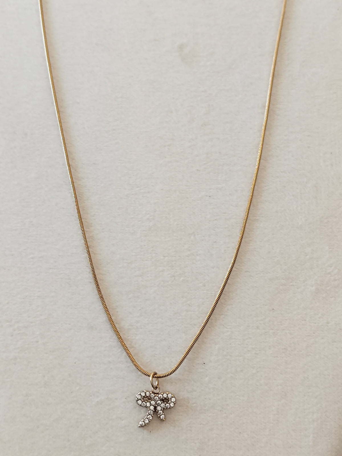 A delicate gold chain necklace with a small bow-shaped pendant adorned with clear rhinestones, displayed on a textured off-white background.