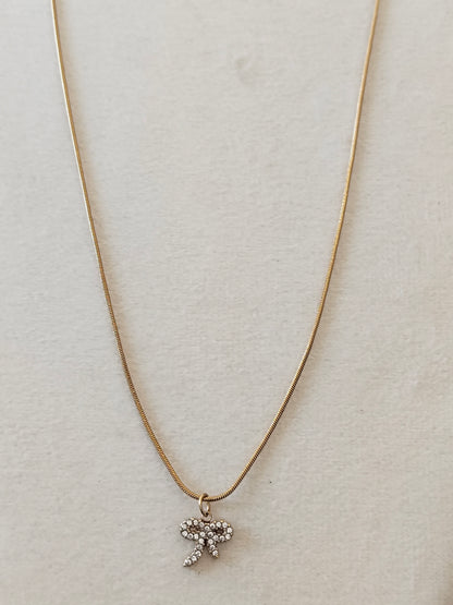 A delicate gold chain necklace with a small bow-shaped pendant adorned with clear rhinestones, displayed on a textured off-white background.