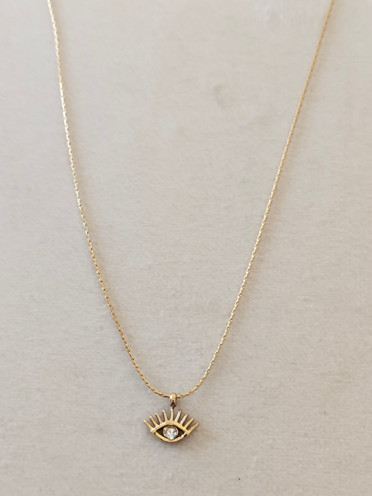 A delicate gold chain necklace featuring a small gold eye pendant with a clear rhinestone, displayed on a textured light beige background.