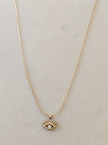 A delicate gold chain necklace featuring a small gold eye pendant with a clear rhinestone, displayed on a textured light beige background.