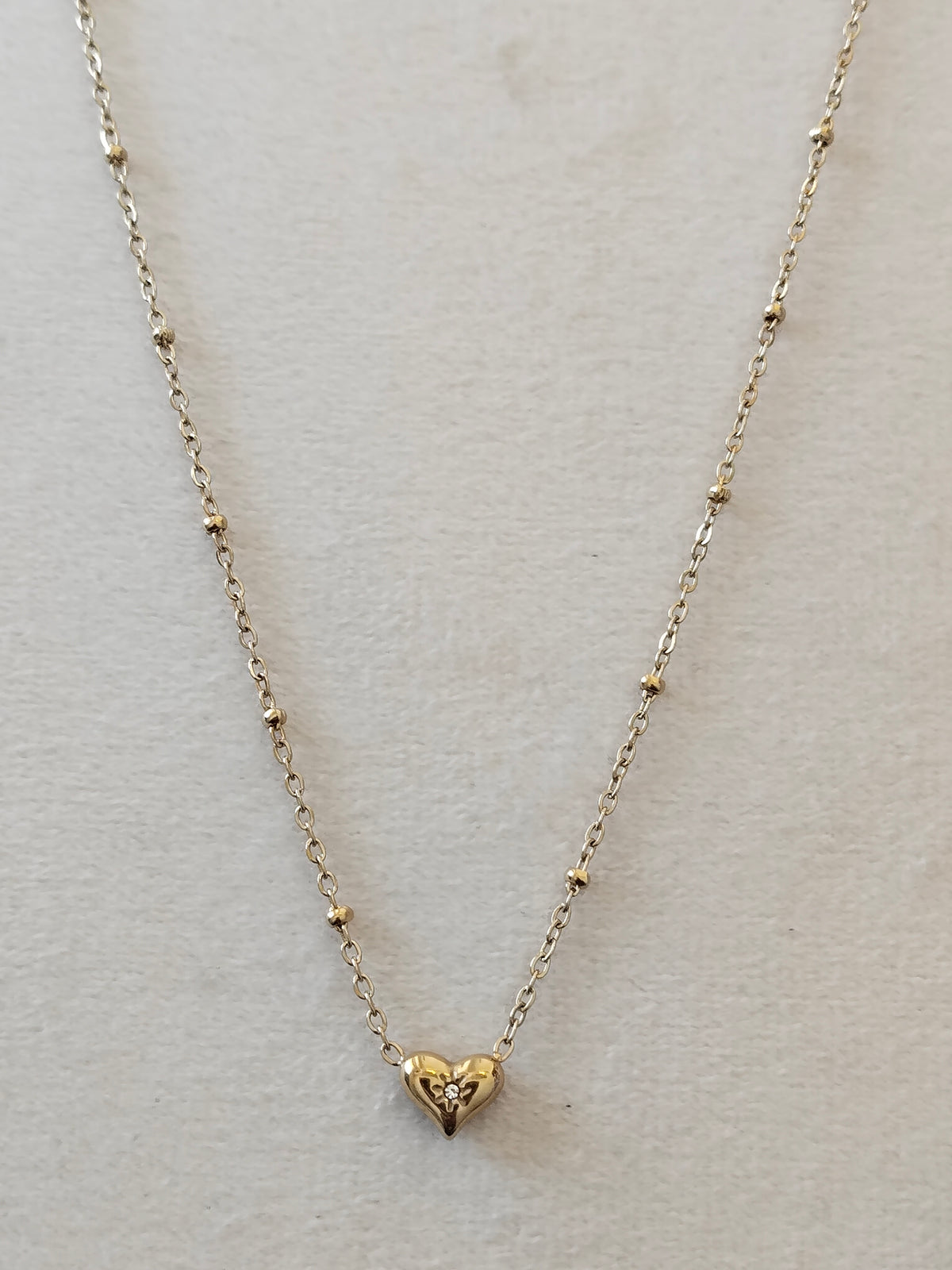 A delicate gold necklace with a small heart-shaped pendant, displayed on a textured off-white background.
