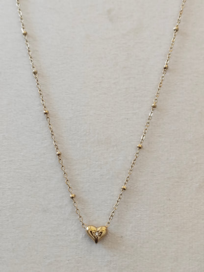 A delicate gold necklace with a small heart-shaped pendant, displayed on a textured off-white background.
