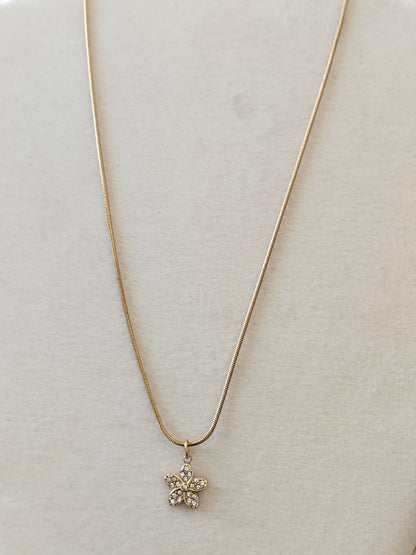 A delicate gold snake chain necklace with a small flower-shaped pendant set with clear rhinestones, displayed on a textured beige surface.