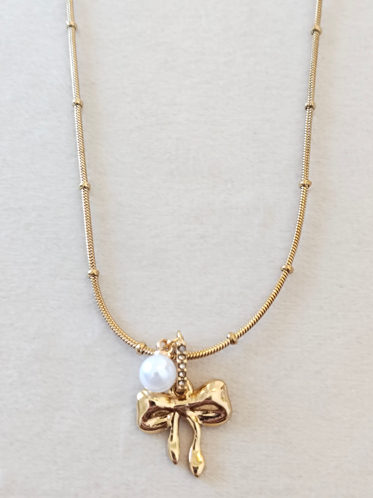 A gold necklace featuring a textured chain with small bead accents, a polished gold bow-shaped pendant, and a small white pearl charm, displayed on a neutral light beige background.