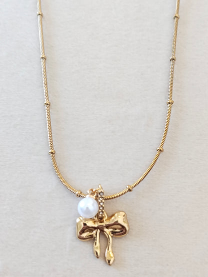 A gold necklace featuring a textured chain with small bead accents, a polished gold bow-shaped pendant, and a small white pearl charm, displayed on a neutral light beige background.